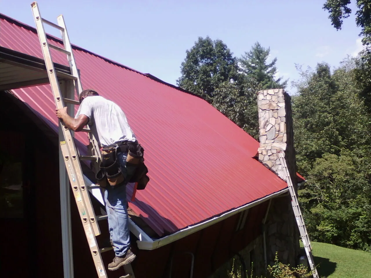 Roofer on a ladder beside a red metal roof and stone chimney — Soffit & Fascia Repair by Roofing Brothers in Yutan